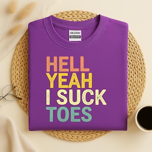 Puede incluir: Una camiseta morada con el texto "HELL YEAH I SUCK TOES" en naranja, amarillo, verde y azul. La camiseta es de algod&oacute;n grueso y tiene cuello redondo. La etiqueta de la marca Gildan es visible.