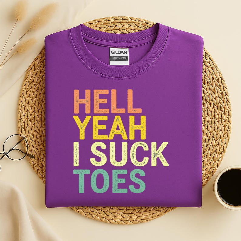 Puede incluir: Una camiseta morada con el texto "HELL YEAH I SUCK TOES" en naranja, amarillo, verde y azul. La camiseta es de algod&oacute;n grueso y tiene cuello redondo. La etiqueta de la marca Gildan es visible.