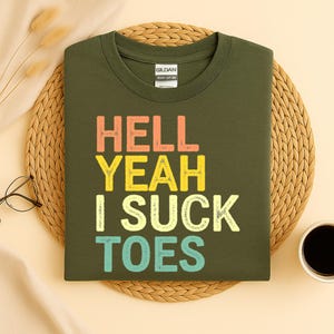 Puede incluir: Una camiseta verde oliva con el texto "HELL YEAH I SUCK TOES" en una fuente retro. La camiseta est&aacute; doblada cuidadosamente sobre una estera tejida, con una taza de caf&eacute; y gafas visibles en el fondo.