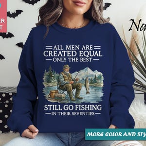 Op de afbeelding: Marineblauwe sweatshirt met de tekst "ALL MEN ARE CREATED EQUAL - ONLY THE BEST - STILL GO FISHING - IN THEIR SEVENTIES." De afbeelding toont een visser met een vis en bergen.