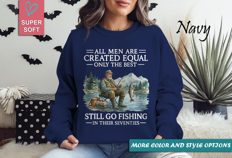 Op de afbeelding: Marineblauwe sweatshirt met de tekst "ALL MEN ARE CREATED EQUAL - ONLY THE BEST - STILL GO FISHING - IN THEIR SEVENTIES." De afbeelding toont een visser met een vis en bergen.