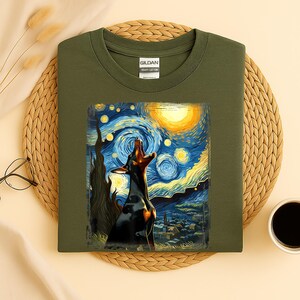 Puede incluir: Camiseta verde oliva con un dise&ntilde;o de un perro negro aullando a la luna, sobre un cielo nocturno arremolinado que recuerda a "La noche estrellada" de Van Gogh. La camiseta es de algod&oacute;n grueso.