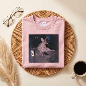 May include: A light pink crew neck t-shirt featuring a graphic print of a hairless cat sitting on a dark surface, holding a lit cigarette in its mouth, with a beverage can and another cigarette nearby. The shirt has a Gildan brand label at the neck.