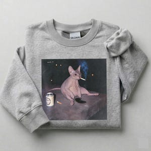 May include: A heather gray crewneck sweatshirt featuring a graphic print of a hairless Sphynx cat sitting on a dark surface. The cat has a cigarette in its mouth, with a silver beverage can and a discarded cigarette butt placed nearby.
