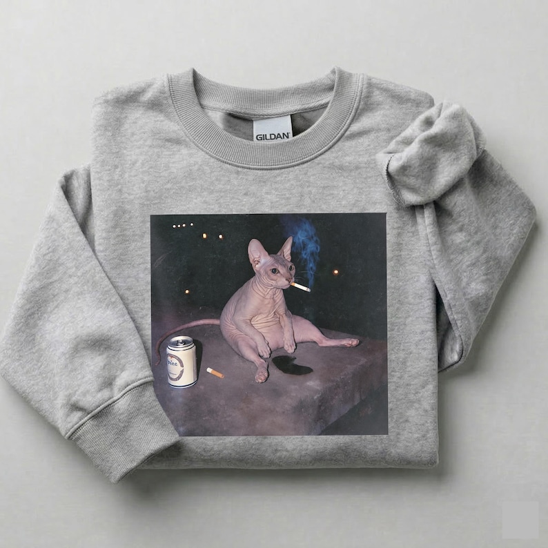 May include: A heather gray crewneck sweatshirt featuring a graphic print of a hairless Sphynx cat sitting on a dark surface. The cat has a cigarette in its mouth, with a silver beverage can and a discarded cigarette butt placed nearby.
