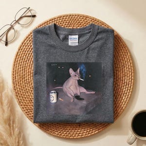May include: A dark gray crew neck t-shirt featuring a graphic print of a hairless cat sitting on a flat surface with a cigarette in its mouth and a small can nearby. The shirt has a Gildan brand label on the inside of the collar and short sleeves.