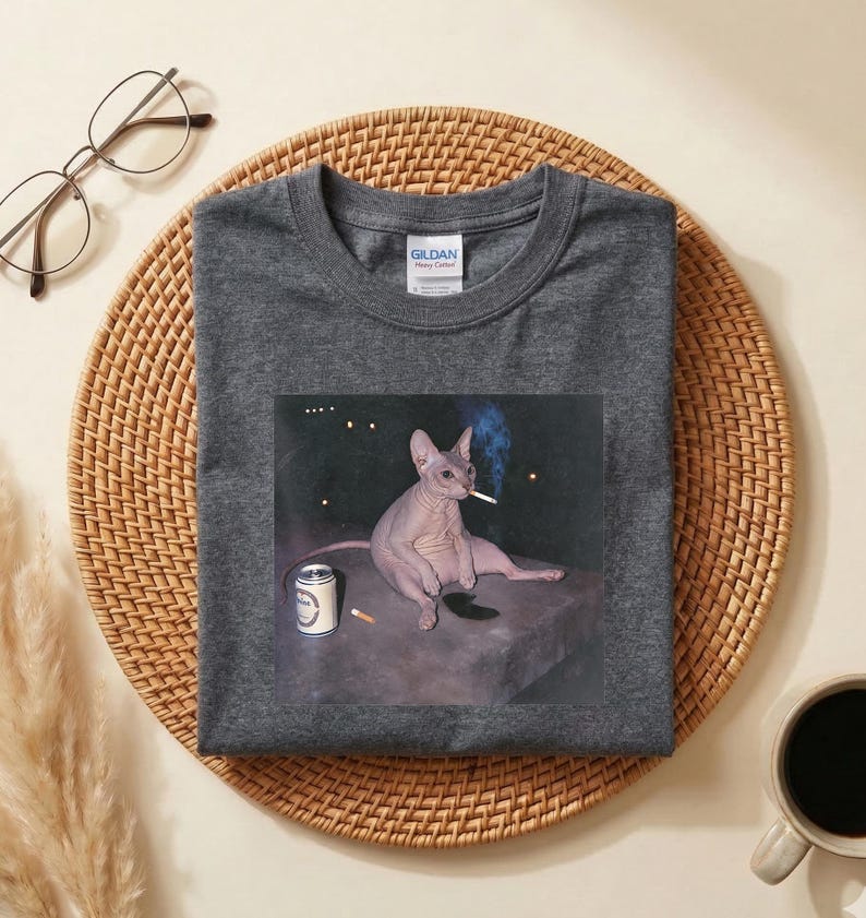 May include: A dark gray crew neck t-shirt featuring a graphic print of a hairless cat sitting on a flat surface with a cigarette in its mouth and a small can nearby. The shirt has a Gildan brand label on the inside of the collar and short sleeves.