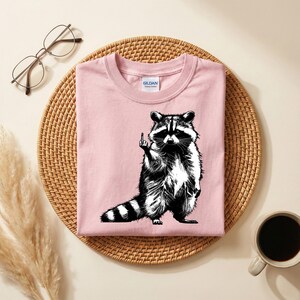 May include: A light pink t-shirt featuring a black and white illustration of a raccoon giving the middle finger. The t-shirt is on a woven, circular placemat, with a pair of glasses and a cup of coffee in the background.