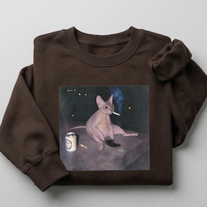 May include: A dark brown crewneck sweatshirt featuring a graphic print of a hairless cat sitting on a ledge, smoking a cigarette with a blue plume of smoke, accompanied by a silver beverage can and a single cigarette lying on the surface.