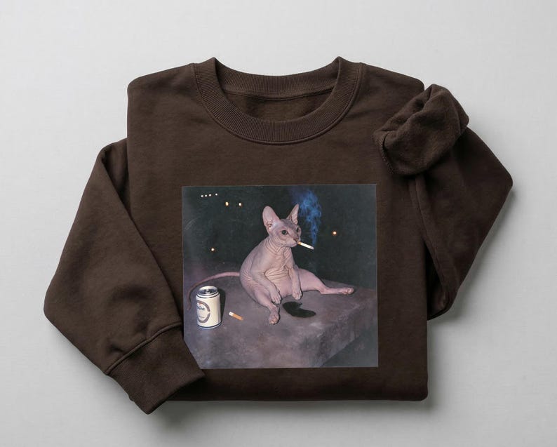 May include: A dark brown crewneck sweatshirt featuring a graphic print of a hairless cat sitting on a ledge, smoking a cigarette with a blue plume of smoke, accompanied by a silver beverage can and a single cigarette lying on the surface.