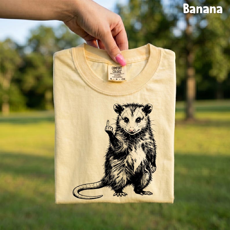 May include: A light yellow t-shirt with a black and white opossum graphic giving the middle finger. The shirt has a crew neck and short sleeves. The text "Banana" is in the upper right corner.