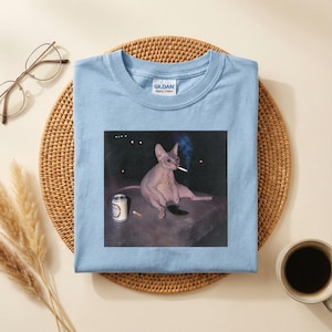 May include: A light blue t-shirt featuring a graphic print of a hairless cat sitting on a dark surface, holding a lit cigarette in its mouth with a small plume of smoke rising, and a silver beverage can placed next to it on the flat surface.