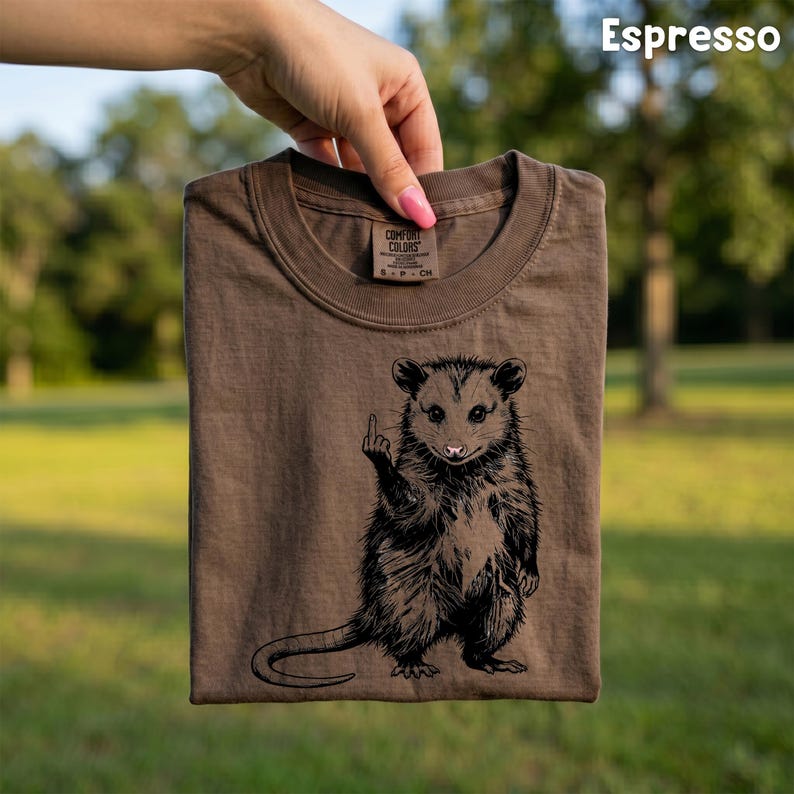 May include: Espresso-colored t-shirt featuring a black graphic of an opossum giving the middle finger. The opossum is standing upright. The shirt is held up by a hand. The shirt has a crew neck and short sleeves. The brand name is visible on the tag.