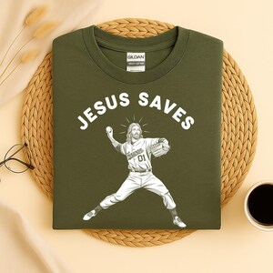 May include: Olive green t-shirt with the text "JESUS SAVES" above a graphic of Jesus in a baseball uniform pitching a ball. The shirt features a crew neck and a tag that reads "GILDAN HEAVY COTTON".