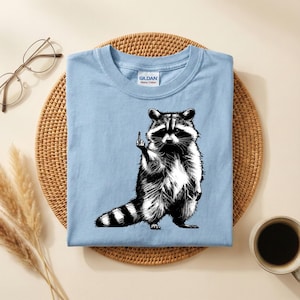 May include: A light blue t-shirt featuring a black and white illustration of a raccoon giving the middle finger. The raccoon is standing upright with its tail striped. The shirt has a crew neck and a tag that says "Gildan Heavy Cotton."