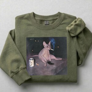 May include: An olive green crewneck sweatshirt featuring a graphic print of a hairless cat sitting on a dark surface. The cat is holding a lit cigarette in its mouth, with a silver beverage can and a second cigarette placed nearby on the surface.
