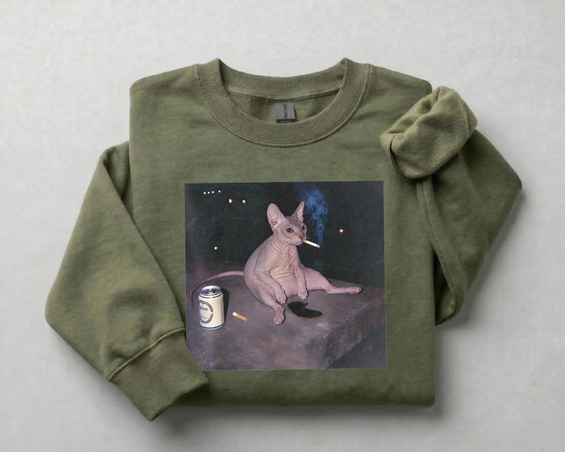 May include: An olive green crewneck sweatshirt featuring a graphic print of a hairless cat sitting on a dark surface. The cat is holding a lit cigarette in its mouth, with a silver beverage can and a second cigarette placed nearby on the surface.