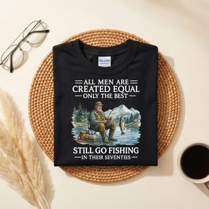 Op de afbeelding: Zwart t-shirt met de tekst "ALL MEN ARE CREATED EQUAL - ONLY THE BEST - STILL GO FISHING - IN THEIR SEVENTIES." Het shirt heeft een afbeelding van een visser die aan een meer zit met bergen op de achtergrond.