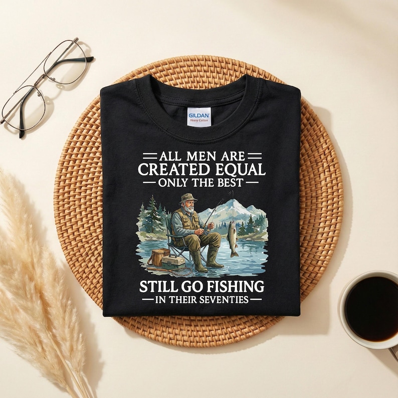 Op de afbeelding: Zwart t-shirt met de tekst "ALL MEN ARE CREATED EQUAL - ONLY THE BEST - STILL GO FISHING - IN THEIR SEVENTIES." Het shirt heeft een afbeelding van een visser die aan een meer zit met bergen op de achtergrond.