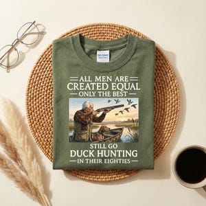 Peut inclure: T-shirt vert olive avec l'inscription "ALL MEN ARE CREATED EQUAL - ONLY THE BEST STILL GO DUCK HUNTING - IN THEIR EIGHTIES." Le motif représente un homme et un chien dans un bateau, chassant des canards. Le t-shirt a un col rond.