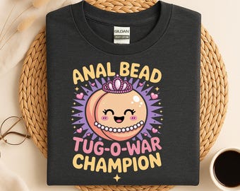 May include: Dark gray t-shirt with the text "ANAL BEAD TUG-O-WAR CHAMPION" and a cartoon graphic of a smiling peach wearing a crown and a pearl necklace. The design includes pink hearts and starbursts.