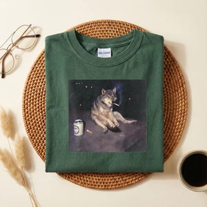 K&ouml;nnte beinhalten: Dunkelgr&uuml;nes T-Shirt mit einem Aufdruck eines Wolfs, der eine Zigarette raucht, mit einer Bierdose und Zigarettenkippen. Der Wolf ist in entspannter Pose vor dunklem Hintergrund dargestellt. Das T-Shirt ist aus weichem Material.