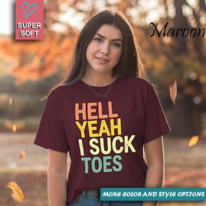 Puede incluir: Camiseta granate con el texto "HELL YEAH I SUCK TOES" en naranja, amarillo y turquesa. La camiseta tambi&eacute;n tiene las palabras "SUPER SOFT" y "MORE COLOR AND STYLE OPTIONS" en la imagen.