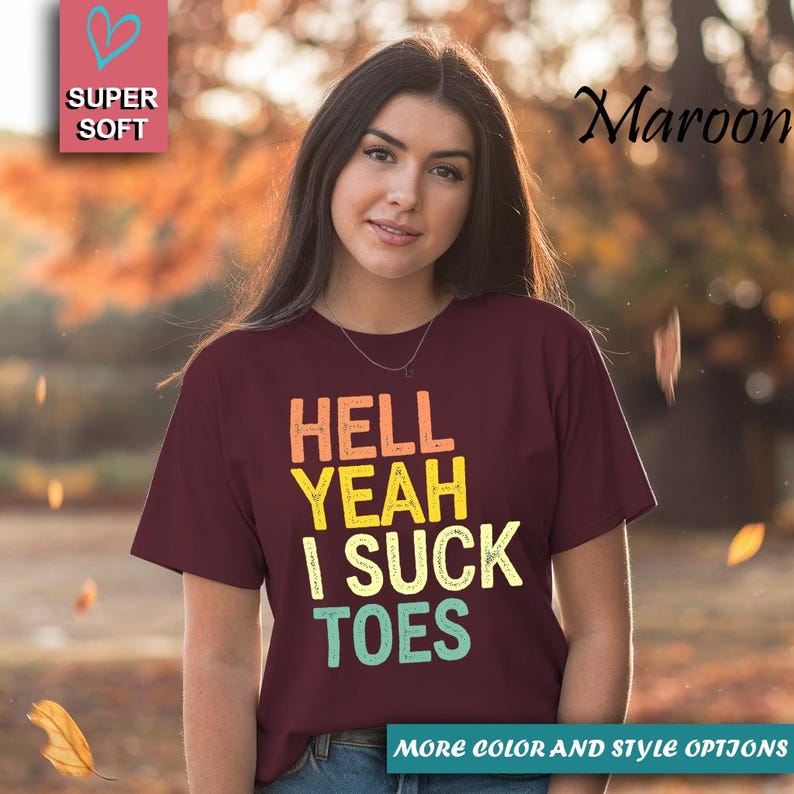 Puede incluir: Camiseta granate con el texto "HELL YEAH I SUCK TOES" en naranja, amarillo y turquesa. La camiseta tambi&eacute;n tiene las palabras "SUPER SOFT" y "MORE COLOR AND STYLE OPTIONS" en la imagen.