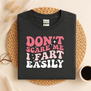 May include: Dark gray t-shirt with the text "DON'T SCARE ME I FART EASILY" in pink and white. The shirt is made of heavy cotton and is displayed on a woven mat.