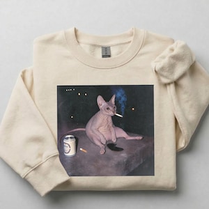 May include: A beige crewneck sweatshirt featuring a graphic print of a hairless cat sitting on a dark surface. The cat is holding a lit cigarette in its mouth with visible smoke, and a silver beverage can sits next to it on the dark surface.