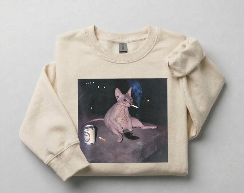 May include: A beige crewneck sweatshirt featuring a graphic print of a hairless cat sitting on a dark surface. The cat is holding a lit cigarette in its mouth with visible smoke, and a silver beverage can sits next to it on the dark surface.