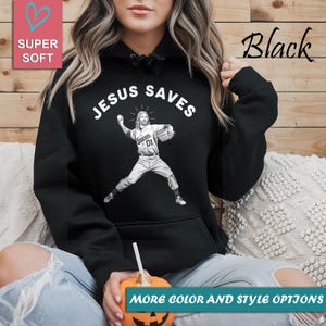 May include: Black hoodie with the text "JESUS SAVES" above a graphic of Jesus pitching a baseball. The hoodie also has the text "Heaven 01" on the jersey. The words "SUPER SOFT" and "MORE COLOR AND STYLE OPTIONS" are also visible.