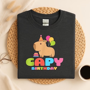May include: Dark gray t-shirt featuring a cartoon capybara wearing a party hat and holding balloons. The shirt has the words "CAPY BIRTHDAY" in colorful letters. The shirt is made of heavy cotton.