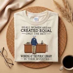 May include: Cream-colored t-shirt with the text "ALL WOMEN ARE CREATED EQUAL - ONLY THE BEST - WORKING AT THE PUBLIC LIBRARY - IN THEIR SEVENTIES." The graphic shows a librarian standing behind a desk. The shirt is on a woven mat.