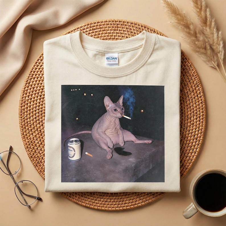 May include: A cream-colored t-shirt featuring a graphic print of a hairless cat sitting on a dark surface, holding a lit cigarette in its mouth with a trail of blue smoke, accompanied by a silver beverage can and a single cigarette on the surface.