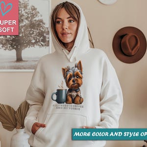 May include: A cream-colored hoodie featuring a graphic of a Yorkshire Terrier with a coffee cup. The text "ALL I NEED IS COFFEE AND MY YORKIE" is printed below the image. The hoodie has a hood and a front pocket.