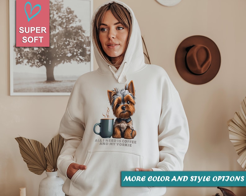 May include: A cream-colored hoodie featuring a graphic of a Yorkshire Terrier with a coffee cup. The text "ALL I NEED IS COFFEE AND MY YORKIE" is printed below the image. The hoodie has a hood and a front pocket.