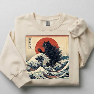 May include: Cream-colored sweatshirt featuring a graphic of a black cat resembling Godzilla, set against a red sun and ocean waves. The design is inspired by Japanese art, with Japanese text. The sweatshirt has a crew neck and long sleeves.