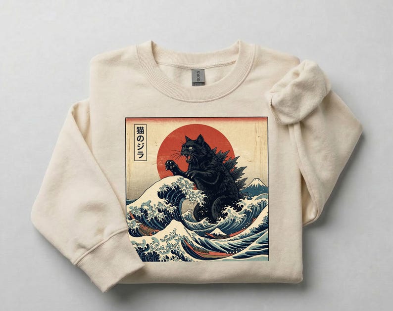 May include: Cream-colored sweatshirt featuring a graphic of a black cat resembling Godzilla, set against a red sun and ocean waves. The design is inspired by Japanese art, with Japanese text. The sweatshirt has a crew neck and long sleeves.
