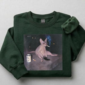 May include: A dark forest green crewneck sweatshirt featuring a graphic print of a hairless cat sitting on a dark surface. The cat is holding a lit cigarette in its mouth, with a silver beverage can and a loose cigarette placed nearby on the surface.