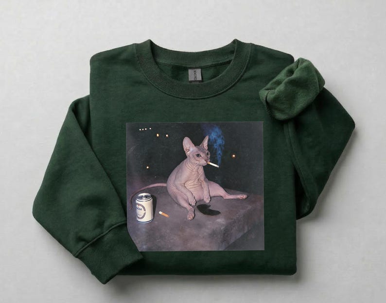 May include: A dark forest green crewneck sweatshirt featuring a graphic print of a hairless cat sitting on a dark surface. The cat is holding a lit cigarette in its mouth, with a silver beverage can and a loose cigarette placed nearby on the surface.