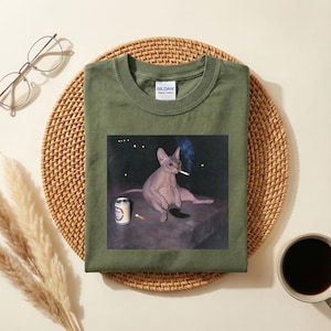 May include: An olive green crew neck t-shirt featuring a graphic print of a hairless Sphynx cat sitting on a dark surface. The cat is holding a lit cigarette in its mouth, with a silver beverage can and another cigarette placed nearby on the surface.