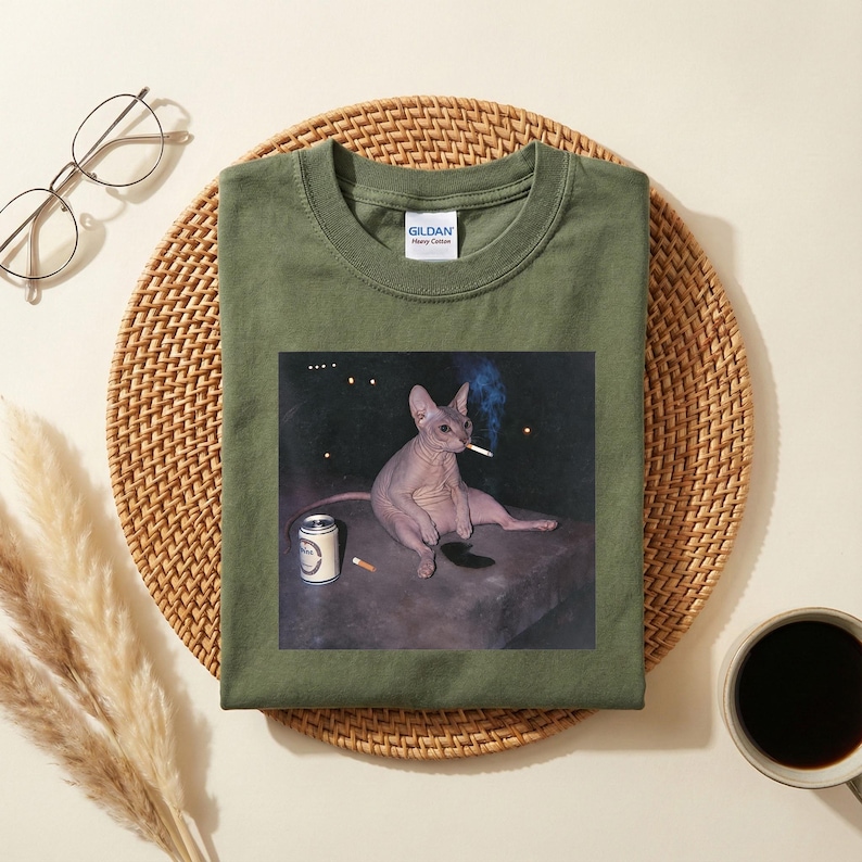 May include: An olive green crew neck t-shirt featuring a graphic print of a hairless Sphynx cat sitting on a dark surface. The cat is holding a lit cigarette in its mouth, with a silver beverage can and another cigarette placed nearby on the surface.