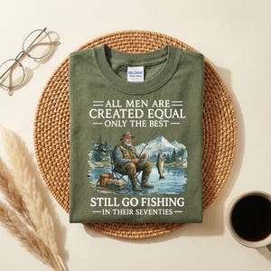 Op de afbeelding: Olijfgroen t-shirt met de tekst "ALL MEN ARE CREATED EQUAL - ONLY THE BEST - STILL GO FISHING - IN THEIR SEVENTIES." De afbeelding toont een visser die aan het vissen is in een meer met bergen op de achtergrond.