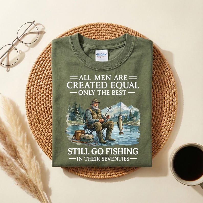 Op de afbeelding: Olijfgroen t-shirt met de tekst "ALL MEN ARE CREATED EQUAL - ONLY THE BEST - STILL GO FISHING - IN THEIR SEVENTIES." De afbeelding toont een visser die aan het vissen is in een meer met bergen op de achtergrond.