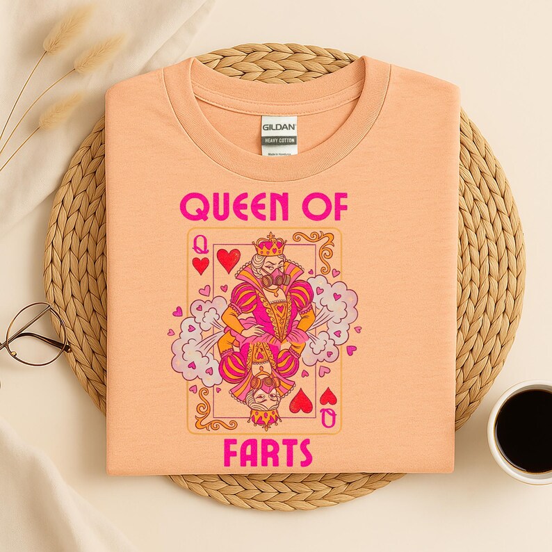 Op de afbeelding: Perzikkleurig T-shirt met een afbeelding van een speelkaart met een koningin met een kroon en gasmasker. De tekst "QUEEN OF FARTS" is in roze gedrukt. Het shirt is gemaakt van zwaar katoen.