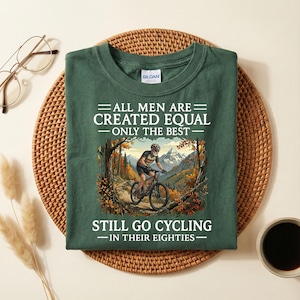 May include: A dark green t-shirt with the text "ALL MEN ARE CREATED EQUAL - ONLY THE BEST - STILL GO CYCLING - IN THEIR EIGHTIES." The graphic shows a cyclist on a mountain bike on a trail with a mountain backdrop.