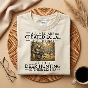 Könnte beinhalten: Cremefarbenes T-Shirt mit dem Text "ALL MEN ARE CREATED EQUAL - ONLY THE BEST - STILL GO DEER HUNTING - IN THEIR SIXTIES." Das Shirt zeigt eine Grafik eines Mannes in Tarnkleidung mit einem Gewehr und einem Hirsch im Hintergrund.