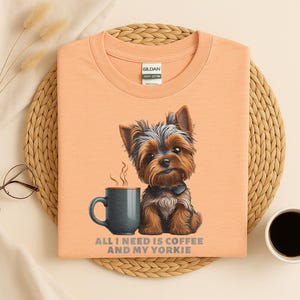 May include: Peach-colored t-shirt featuring a Yorkshire Terrier illustration with a coffee mug. The text "ALL I NEED IS COFFEE AND MY YORKIE" is printed below the image. The shirt is made of heavy cotton.
