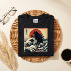 May include: Black t-shirt featuring a graphic of a black cat in a Godzilla-like pose, set against a red sun and ocean waves. The design is inspired by Japanese art, with text in Japanese. The shirt is on a woven mat.